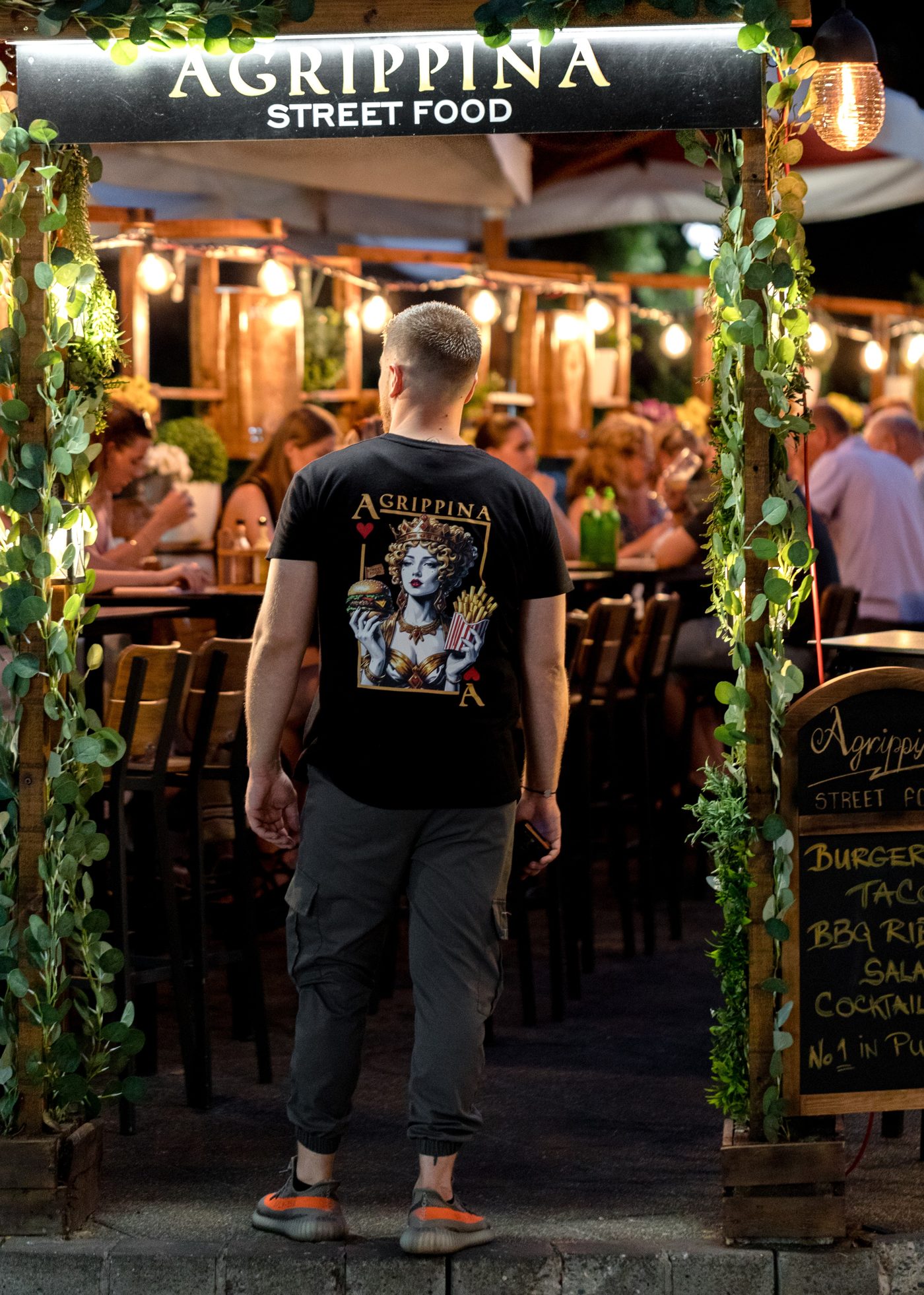 Gost u Agrippina majici s kraljicom iz igraće karte stoji pod osvijetljenim lukom Agrippina Street Food; iza njega puna terasa i ploča s ponudom